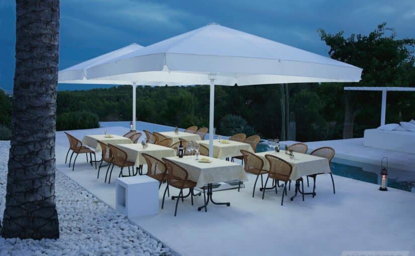 To store hvide parasoller over spiseborde og stole på en lys terrasse ved en swimmingpool.